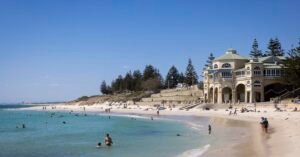 People enjoying the sun and swimming at Cottesloe Beach in Perth, Australia, with the iconic Indiana Tea House building in the background.