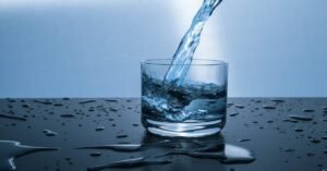 A stream of water being poured into a clear glass on a dark surface with water droplets scattered around.