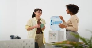 Two women in an office having a conversation while standing near a water cooler, each holding a mug.