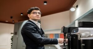 A man in a suit and glasses using a coffee machine in an office setting, looking slightly to the side.